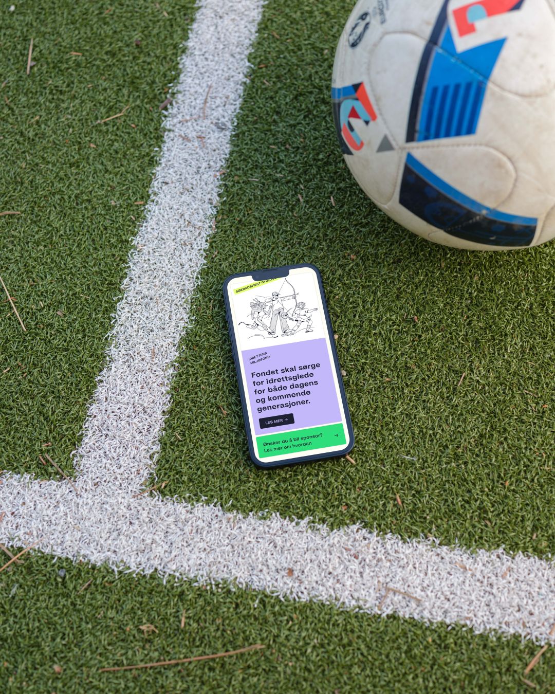 En telefon med grontlag.no åpen står på en fotballbane ved siden av en ball.