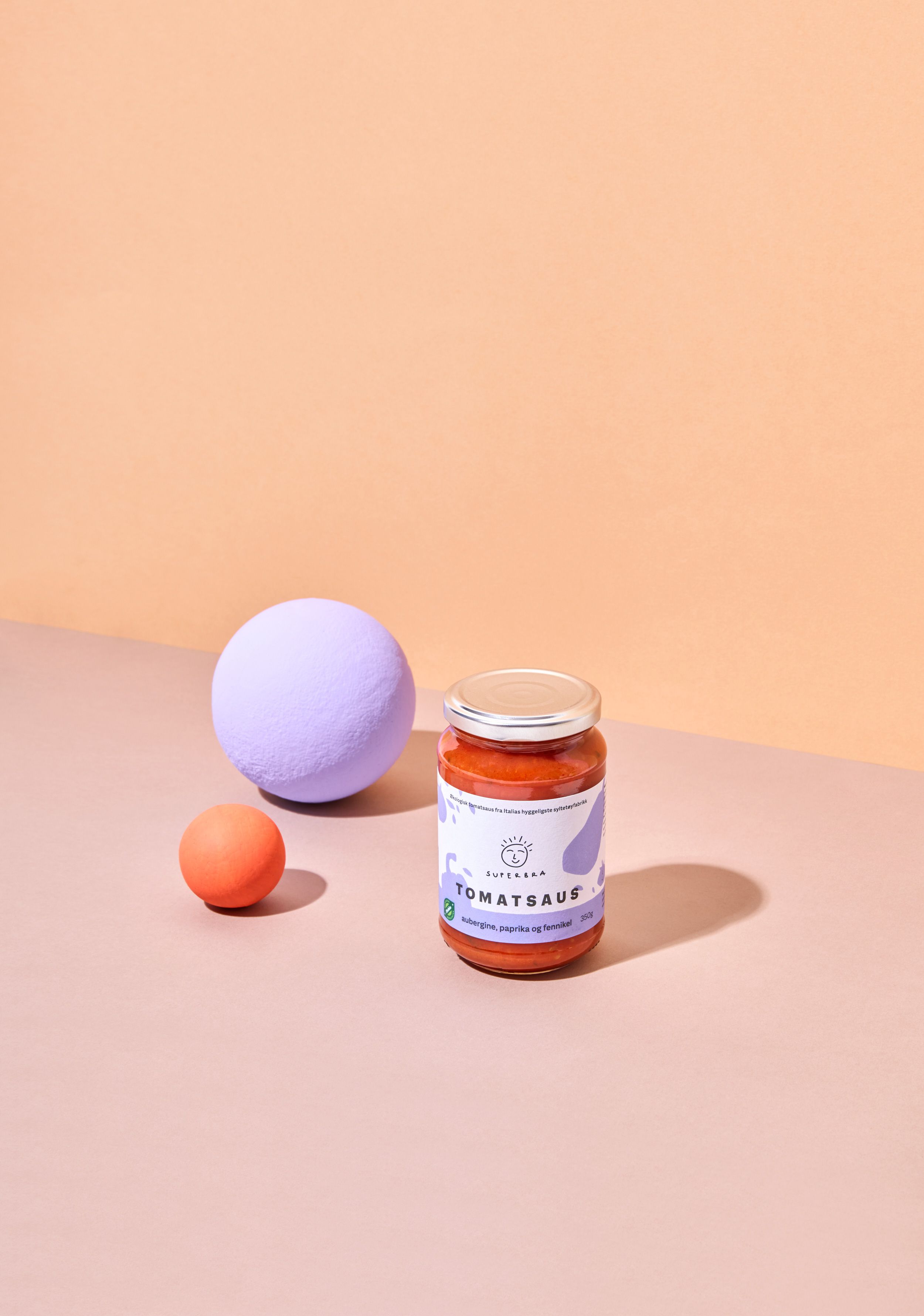 Super Bra tomatsaus, på oransj bord forran en oransj vegg, med to baller i oransj og lilla til sin venstre.